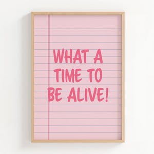 May include: A framed print with the phrase "WHAT A TIME TO BE ALIVE!" in pink, handwritten-style font. The background is a light pink color with horizontal lines, resembling notebook paper. The frame is a light wood color.