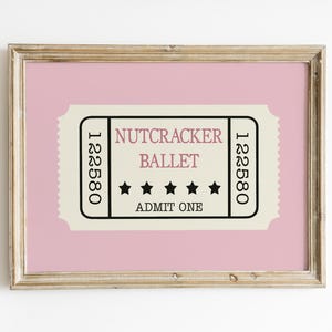 May include: A framed print featuring a pink background with a vintage-style ticket design. The ticket reads "Nutcracker Ballet" with stars and "Admit One." The frame is a light, natural wood color.