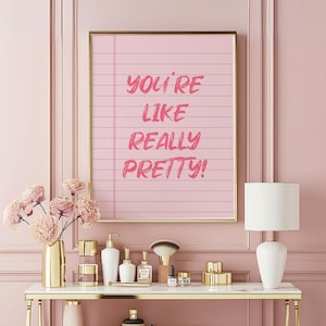 Può includere: Una stampa incorniciata con la scritta "YOU'RE LIKE REALLY PRETTY!" in rosa su uno sfondo di carta a righe rosa. La stampa con cornice dorata è appesa sopra un tavolo bianco e oro con prodotti di bellezza e un vaso d'oro con fiori rosa.