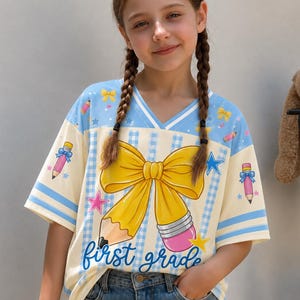 Könnte beinhalten: Ein hellblaues und cremefarbenes T-Shirt mit V-Ausschnitt, großem gelben Schleifenmotiv, rosa Stiften, blauen Sternen und dem Schriftzug first grade in blauer Schreibschrift. Das Shirt hat ein hellblau-cremefarbenes Karomuster und blaue Ärmelstreifen.