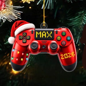 May include: A red video game controller ornament featuring a Santa hat, a string of colourful lights, and the text MAX and 2026 in yellow pixelated font. The controller includes icons of a tree, gift, snowflake, and Santa face on the buttons.
