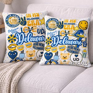 Op de afbeelding: Twee decoratieve sierkussens met een witte achtergrond en een collage van Delaware-thema graphics. De kussens bevatten tekst als "Delaware Vibes", "Blue Hens" en "In My Delaware Era", samen met verschillende illustraties in blauw, geel en wit.