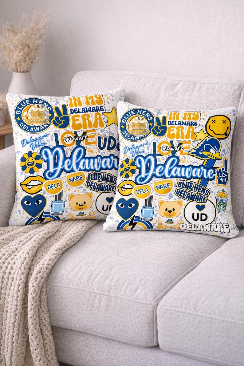 Op de afbeelding: Twee decoratieve sierkussens met een witte achtergrond en een collage van Delaware-thema graphics. De kussens bevatten tekst als "Delaware Vibes", "Blue Hens" en "In My Delaware Era", samen met verschillende illustraties in blauw, geel en wit.