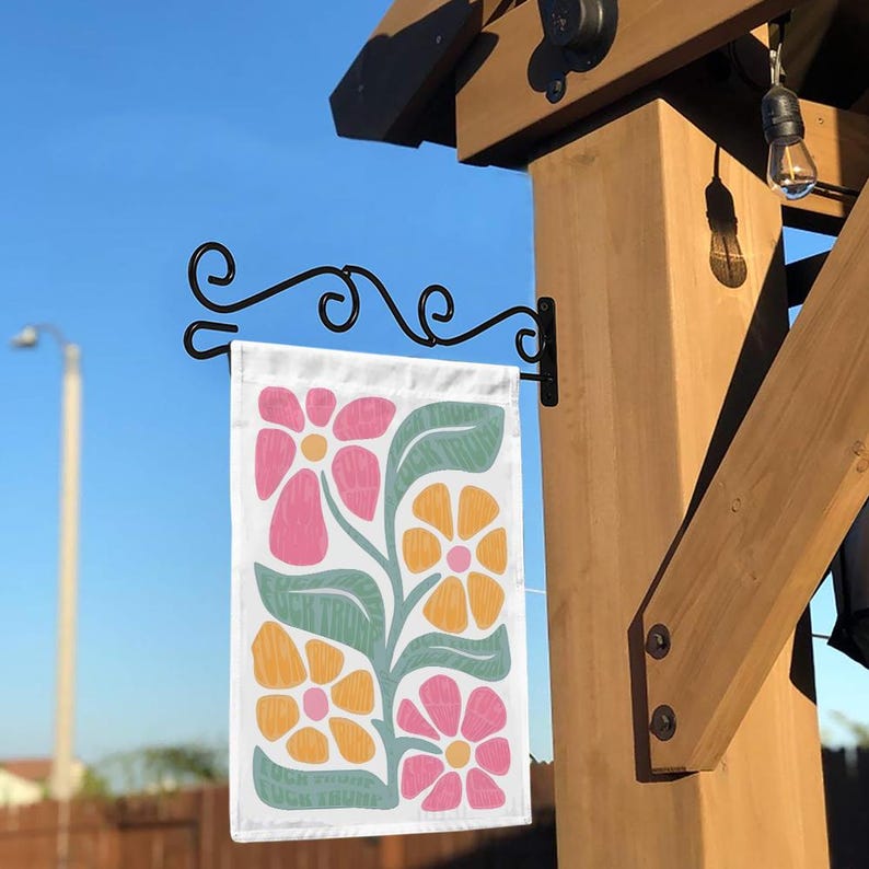 Puede incluir: Una bandera de jard&iacute;n blanca con un dise&ntilde;o floral. La bandera presenta flores rosas, amarillas y verdes sobre un fondo blanco. La bandera est&aacute; colgada de un asta de bandera de metal negro.