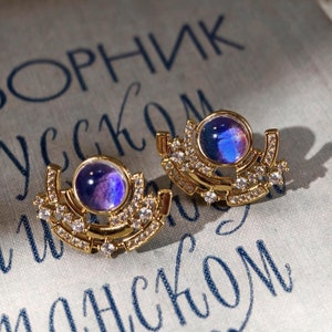 May include: A pair of gold earrings with a purple iridescent stone set in each. The earrings are shaped like a crescent moon with a row of small clear stones around the edge.