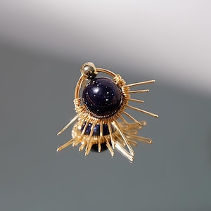 Puede incluir: Un pendiente de alambre dorado con una esfera de piedra preciosa azul oscuro en el centro. El alambre está formado en un diseño de sol radiante con múltiples puntos que irradian hacia afuera.