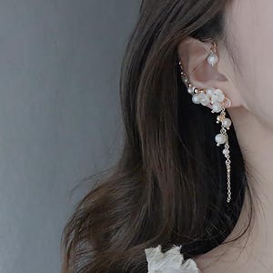 May include: A gold-toned ear cuff and earring set featuring white pearl accents. The ear cuff curves along the ear, adorned with pearls and floral details. The earring dangles with a chain of pearls, creating an elegant, nature-inspired design.