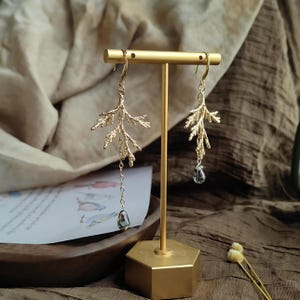 The gold pine branch earrings displayed on a modern, hexagonal brass stand in an indoor setting. The soft, warm light highlights the artistic, sculptural quality of the whimsical, asymmetrical jewelry design.
