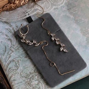 Könnte beinhalten: Vergoldetes Ohr-Manschetten- und Ohrring-Set. Die Ohr-Manschette und die Ohrringe weisen ein zartes Blattdesign auf, das mit klaren Kristallen verziert ist. Die Ohrringe haben ein langes Ketten-Design. Der Schmuck wird auf einer grauen Samtoberfläche präsentiert.