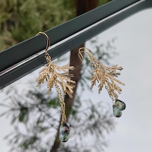 The asymmetrical pine branch earrings shown artistically reflected in a mirror against a blurred, natural outdoor background. This creative angle showcases both the front and a partial side view of the intricate botanical design.