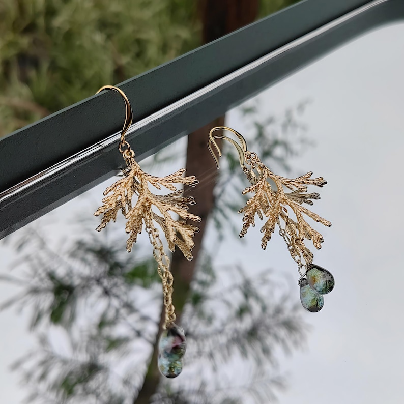 The asymmetrical pine branch earrings shown artistically reflected in a mirror against a blurred, natural outdoor background. This creative angle showcases both the front and a partial side view of the intricate botanical design.