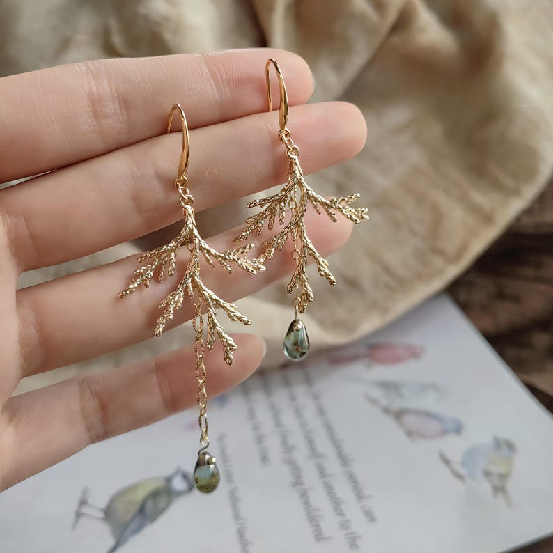 A person's hand holding the pair of asymmetrical pine branch earrings against a soft background with a botanical bird illustration. This image provides a clear sense of the earrings' actual size, scale, and delicate handcrafted details.