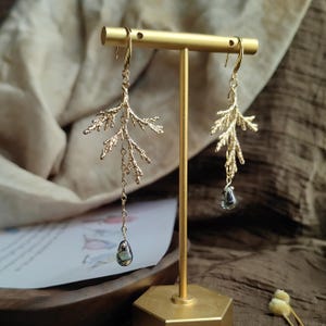 The gold pine branch earrings displayed on a modern, hexagonal brass stand in an indoor setting. The soft, warm light highlights the artistic, sculptural quality of the whimsical, asymmetrical jewelry design.