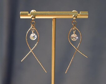 May include: A pair of gold dangle earrings with a clear crystal at the end of each earring. The earrings are hanging from a gold stand.