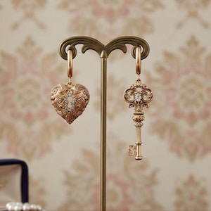 Peut inclure: Boucles d'oreilles dorées, l'une en forme de cœur avec un motif de trou de serrure et l'autre en forme de clé. Les deux boucles d'oreilles présentent des détails complexes et sont ornées de petits cristaux transparents. Elles sont présentées sur un présentoir décoratif.