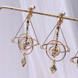 May include: A pair of gold-toned clip-on earrings featuring a geometric pyramid frame surrounding concentric rings and a central sphere. A small, diamond-shaped charm with a tiny embedded stone hangs from the bottom of each earring structure.