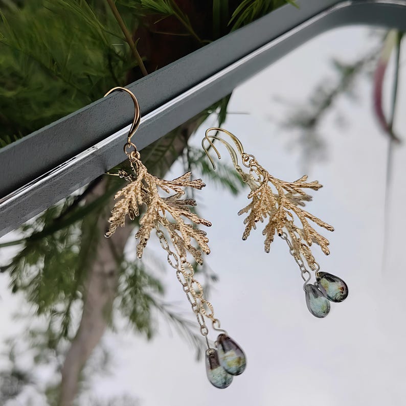 The asymmetrical pine branch earrings shown artistically reflected in a mirror against a blurred, natural outdoor background. This creative angle showcases both the front and a partial side view of the intricate botanical design.