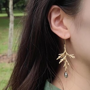 A close-up view of a model's ear wearing the asymmetrical gold pine branch earring. The photo is taken outdoors in a park setting, clearly demonstrating the earring's graceful drop length and scale against the skin.