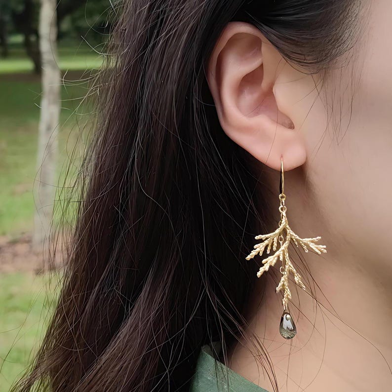A close-up view of a model's ear wearing the asymmetrical gold pine branch earring. The photo is taken outdoors in a park setting, clearly demonstrating the earring's graceful drop length and scale against the skin.
