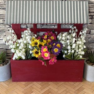 May include: A red wooden planter box with a corrugated metal roof, filled with white lily of the valley, yellow sunflowers, and pink and purple flowers. Two small potted succulents flank the planter.