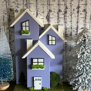 Puede incluir: Una casa decorativa de madera en tonos lavanda, con molduras y chimeneas blancas. La casa presenta ventanas con marcos negros con detalles verdes y una pequeña puerta blanca. Dos árboles de Navidad en miniatura flanquean la casa.