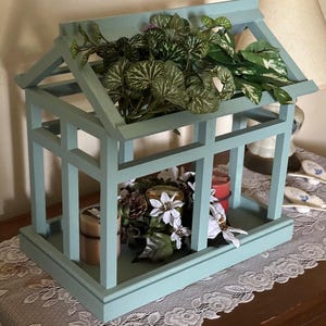 Könnte beinhalten: Ein hellblauer Kerzenhalter aus Holz mit einem dekorativen Dach und zwei Ablagen. Der Halter ist mit Grünpflanzen und Kerzen dekoriert.