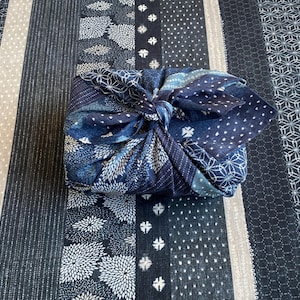 Può includere: Un oggetto quadrato avvolto in un tessuto blu scuro con un nodo sulla parte superiore. Il tessuto presenta un patchwork di vari motivi, tra cui disegni floreali bianchi, forme geometriche e piccoli punti, poggiato su una superficie tessile a righe.