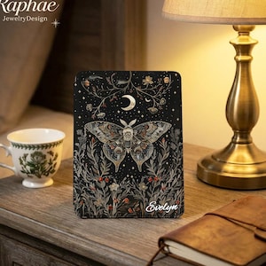 May include: A black e-reader cover featuring a central moth illustration, a crescent moon, and botanical elements in gold and muted tones. The name Evelyn is printed in white script at the bottom right corner of the rectangular protective case.