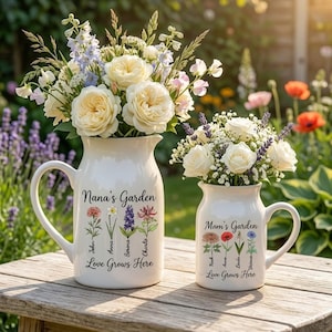 Peut inclure: Deux pichets en céramique blanche avec des compositions florales. Le plus grand affiche "Nana's Garden" avec des noms et des illustrations de fleurs. Le plus petit affiche "Mom's Garden" avec des noms et des illustrations de fleurs. Les deux pichets ont l'inscription "Love Grows Here".