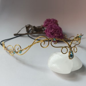 K&ouml;nnte beinhalten: Ein Diadem aus goldfarbenem Draht mit kunstvollen, geschwungenen Metallmustern und kleinen hellblauen Kristallen. Ein gr&ouml;&szlig;erer, tropfenf&ouml;rmiger blauer Kristall h&auml;ngt in der Mitte des Kopfschmucks, der mit einer d&uuml;nnen schwarzen Kordel befestigt ist.
