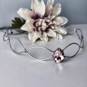 Puede incluir: Una corona de alambre plateado con un diseño ovalado y una gema rosa en forma de lágrima. Una flor grande y clara con un centro oscuro en el fondo. La corona es para una boda o una ocasión especial.