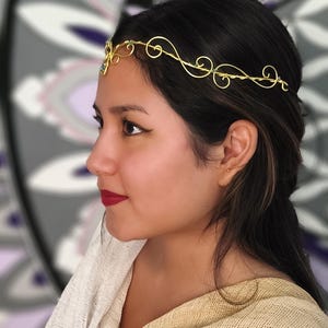 K&ouml;nnte beinhalten: Ein Kopfschmuck aus goldfarbenem Draht mit zarten Wirbelmustern und kleinen gr&uuml;nen Akzenten. Das Accessoire ist so gestaltet, dass es auf der Stirn getragen wird, mit einem zentralen Blickfang und d&uuml;nnen, flexiblen Drahtb&auml;ndern an den Seiten.