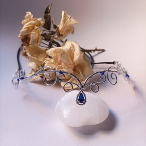 K&ouml;nnte beinhalten: Ein silberfarbener Draht-Kopfschmuck mit komplexen, geschwungenen Metallmustern, kleinen blauen Perlen und einem zentralen tropfenf&ouml;rmigen blauen Edelsteinanh&auml;nger. Das St&uuml;ck besteht aus d&uuml;nnem, flexiblem Draht in symmetrischen Schlaufen.