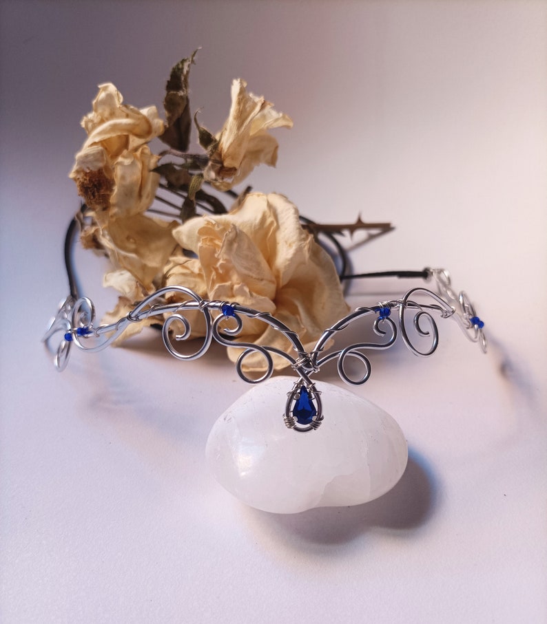 K&ouml;nnte beinhalten: Ein silberfarbener Draht-Kopfschmuck mit komplexen, geschwungenen Metallmustern, kleinen blauen Perlen und einem zentralen tropfenf&ouml;rmigen blauen Edelsteinanh&auml;nger. Das St&uuml;ck besteht aus d&uuml;nnem, flexiblem Draht in symmetrischen Schlaufen.