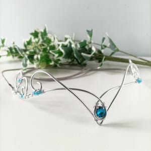 Puede incluir: Diadema de alambre plateado con una piedra preciosa azul y cristales azules más pequeños. La diadema está diseñada en un estilo delicado y fluido.