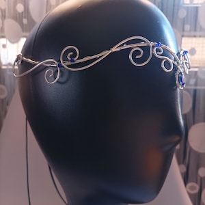 K&ouml;nnte beinhalten: Ein silberfarbener Draht-Kopfschmuck mit kunstvollen, geschwungenen Metallmustern und kleinen, leuchtend blauen Perlen. Das Stirnband ist so gestaltet, dass es auf der Stirn aufliegt und mit einer d&uuml;nnen schwarzen Kordel am Hinterkopf befestigt wird.