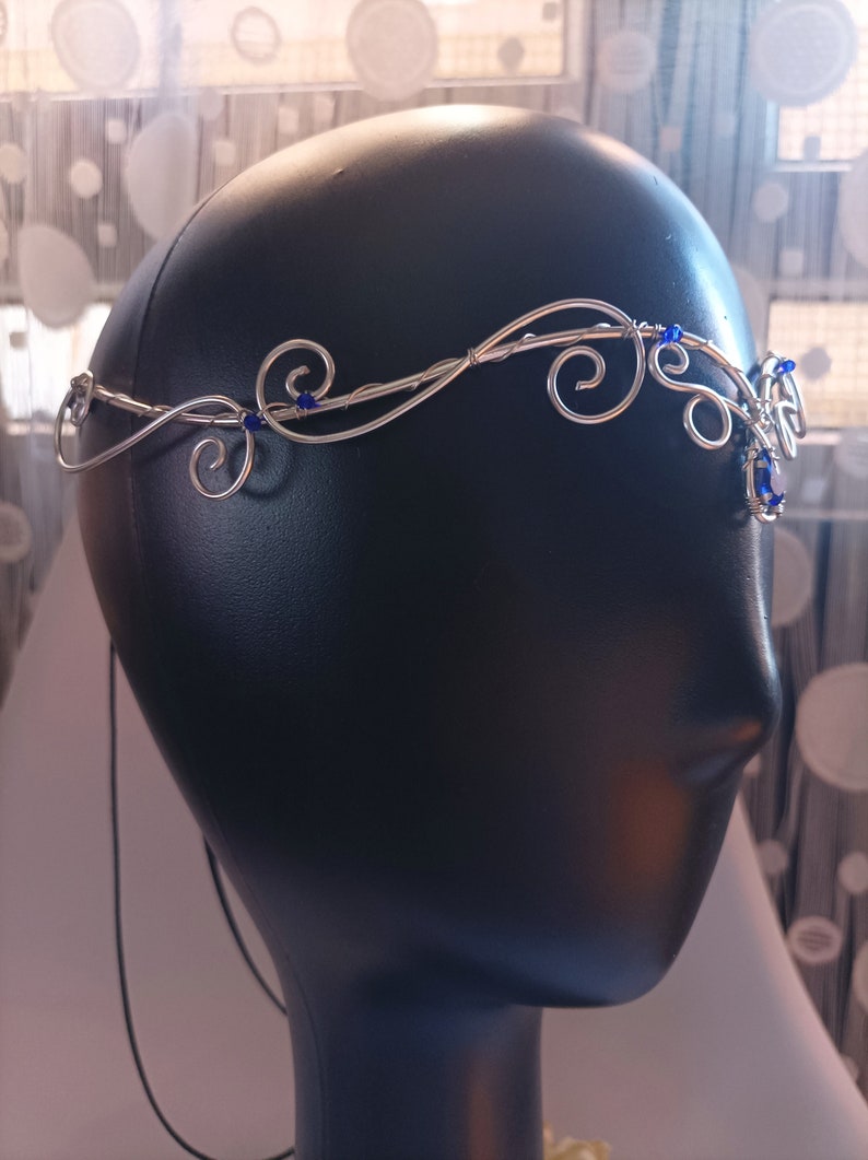 K&ouml;nnte beinhalten: Ein silberfarbener Draht-Kopfschmuck mit kunstvollen, geschwungenen Metallmustern und kleinen, leuchtend blauen Perlen. Das Stirnband ist so gestaltet, dass es auf der Stirn aufliegt und mit einer d&uuml;nnen schwarzen Kordel am Hinterkopf befestigt wird.