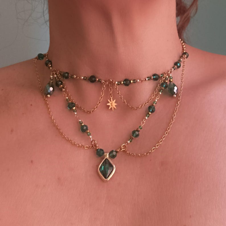 May include: A gold layered necklace with a green gemstone pendant and a star charm. The necklace is made of small beads and chains.