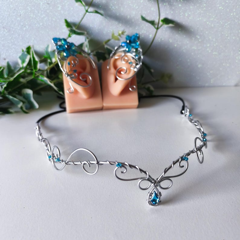 K&ouml;nnte beinhalten: Ein silbernes Draht-Stirnband mit geschwungenen Metallmustern und kleinen blauen Perlen, begleitet von einem Paar passender silberner Draht-Ohrklemmen, die mit blauen floralen Akzenten und Perlen verziert sind, f&uuml;r Ohren und Stirn.