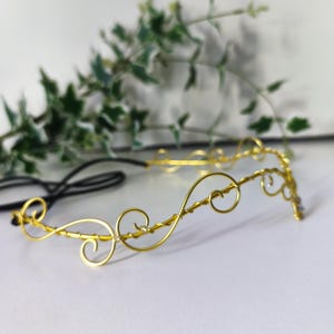 K&ouml;nnte beinhalten: Ein goldfarbenes Draht-Stirnband mit einer Reihe von miteinander verbundenen, geschwungenen Schlaufen und Kurven. Der Metalldraht ist zu einem durchgehenden Wellenmuster geformt, in das kleine, klare, reflektierende Perlen eingearbeitet sind, befestigt an einem schwarzen Gummiband.