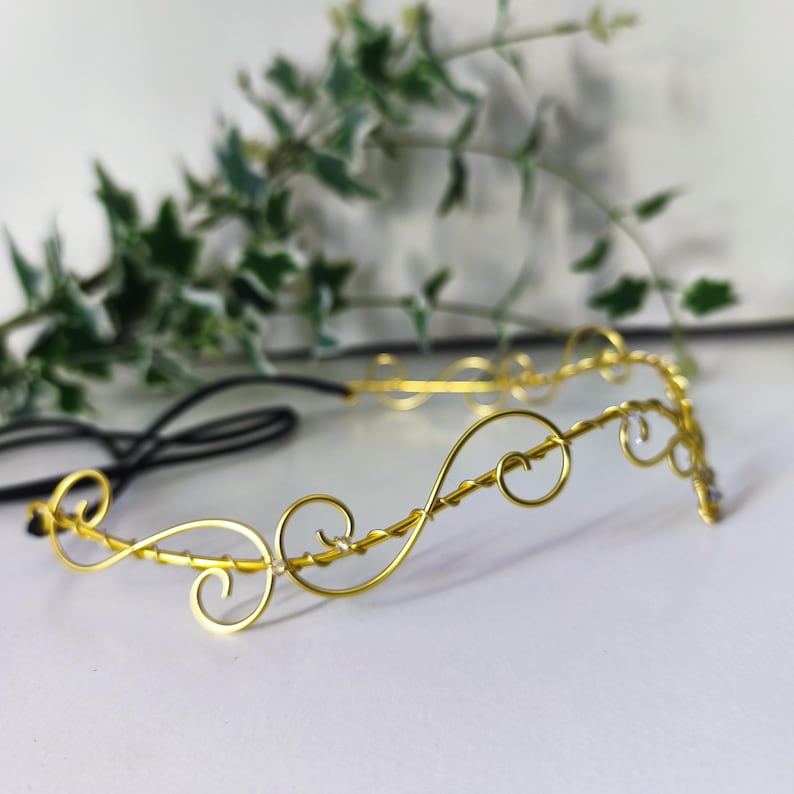 K&ouml;nnte beinhalten: Ein goldfarbenes Draht-Stirnband mit einer Reihe von miteinander verbundenen, geschwungenen Schlaufen und Kurven. Der Metalldraht ist zu einem durchgehenden Wellenmuster geformt, in das kleine, klare, reflektierende Perlen eingearbeitet sind, befestigt an einem schwarzen Gummiband.