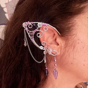 May include: A silver ear cuff featuring a floral and swirl design, embellished with pink and purple beads and dangling charms. The ear cuff is worn on the ear, with the design wrapping around the ear.