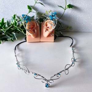 K&ouml;nnte beinhalten: Ein silberfarbenes Draht-Stirnband mit einem zentralen tropfenf&ouml;rmigen blauen Kristall und kleinen blauen Perlenakzenten. Das Set enth&auml;lt zwei passende Ohrklemmen aus Silberdraht, die jeweils mit einer blauen Kristallblume und Drahtspiralen verziert sind.