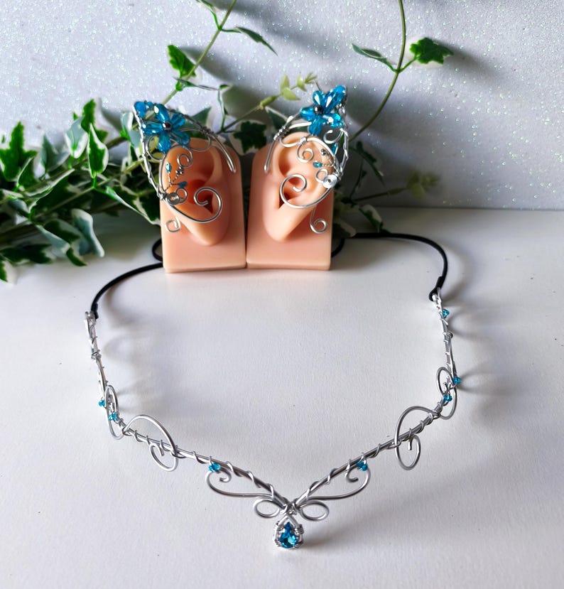 K&ouml;nnte beinhalten: Ein silberfarbenes Draht-Stirnband mit einem zentralen tropfenf&ouml;rmigen blauen Kristall und kleinen blauen Perlenakzenten. Das Set enth&auml;lt zwei passende Ohrklemmen aus Silberdraht, die jeweils mit einer blauen Kristallblume und Drahtspiralen verziert sind.