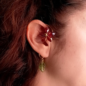 Puede incluir: Un pendiente de aro dorado con un diseño de flor roja y un colgante de hoja verde.