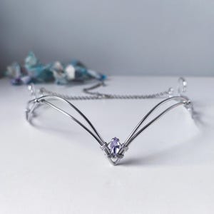 Puede incluir: Una tiara plateada con una gema central de color púrpura claro, con corte marquesa. Esta delicada pieza para la cabeza presenta un diseño de cadena y alambre, perfecta para bodas o eventos de fantasía.