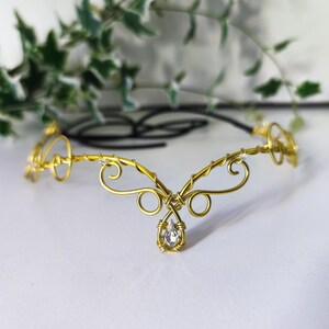 K&ouml;nnte beinhalten: Ein goldfarbener Kopfschmuck aus Draht mit komplizierten, geschwungenen Metallmustern und einem zentralen, tropfenf&ouml;rmigen, klaren Kristallanh&auml;nger. Das Accessoire besteht aus d&uuml;nnem, flexiblem Draht und hat eine schwarze Kordel zur Anpassung.
