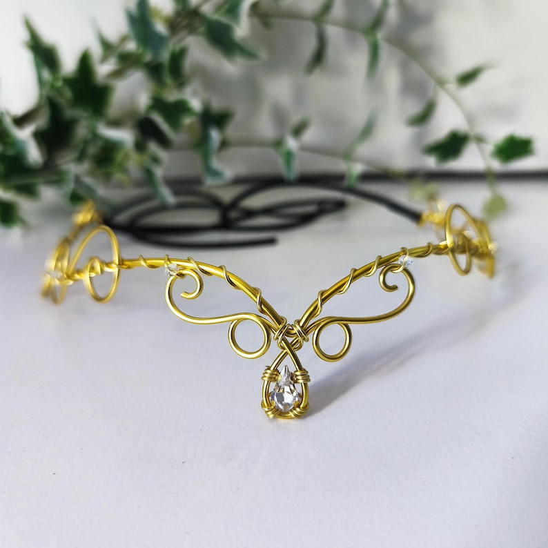 K&ouml;nnte beinhalten: Ein goldfarbener Kopfschmuck aus Draht mit komplizierten, geschwungenen Metallmustern und einem zentralen, tropfenf&ouml;rmigen, klaren Kristallanh&auml;nger. Das Accessoire besteht aus d&uuml;nnem, flexiblem Draht und hat eine schwarze Kordel zur Anpassung.