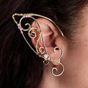 May include: A gold-coloured ear cuff in an elven style. The cuff has intricate wirework, swirls, and small pink beads. The design curves around the ear, creating an elegant, fantasy-inspired accessory.