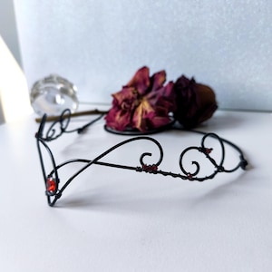 May include: A black wire tiara featuring a pointed centre design with a single round red gemstone at the peak and smaller red accents along the curved wire frame. The headpiece is constructed from thin, dark metal wire shaped into swirls.
